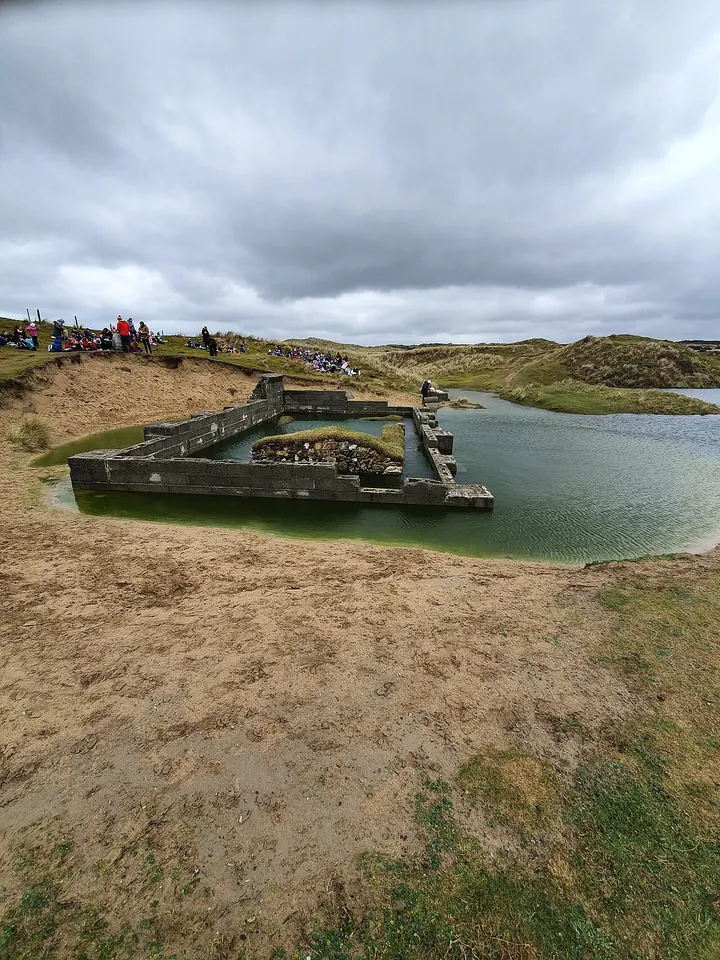 The low stone walls of a small ancient chapel sunk into a sandy hollow among grassy dunes, partly flooded, with people gathered on the dune ridge behind it