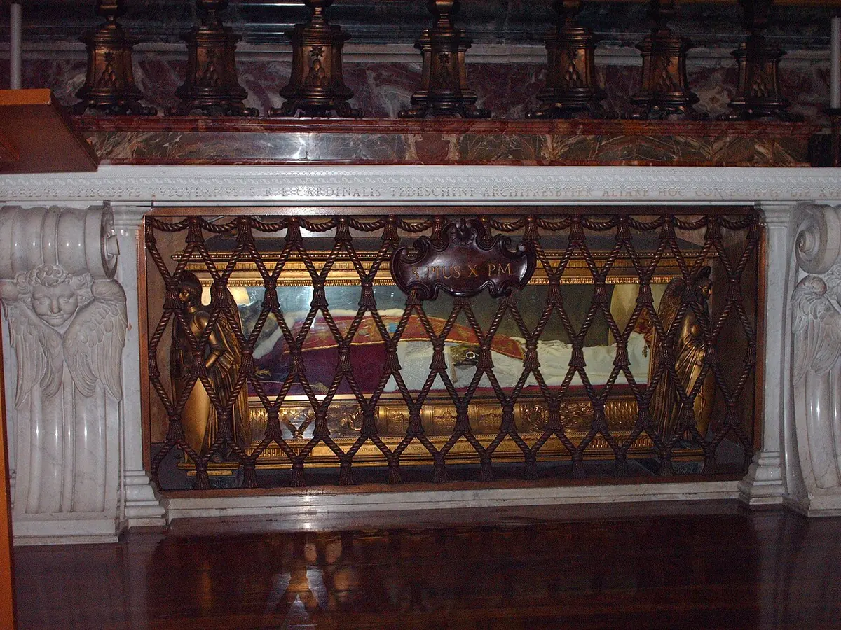 The body of Pius X in red vestments lying in a glass and gilt bronze casket beneath a marble altar lined with tall candlesticks