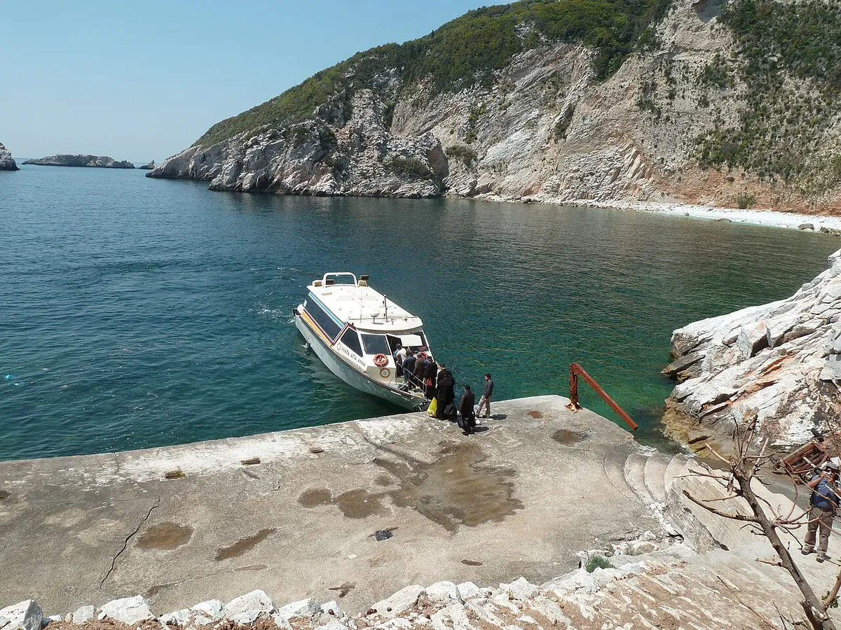A small boat moored at a rocky concrete landing in a clear blue-green bay below steep white cliffs, with monks in black boarding