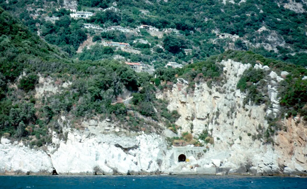 A white domed church and monastery buildings among dense green trees on a steep mountainside, with small houses scattered above
