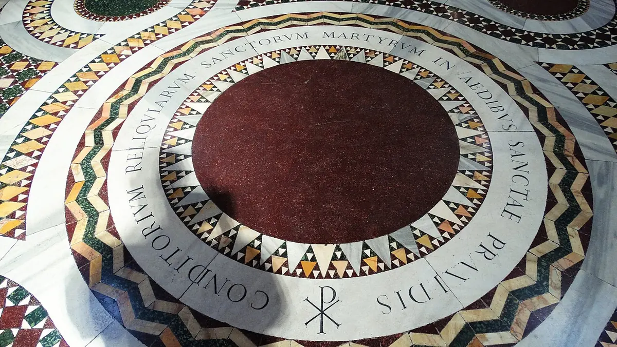 A round slab of dark red porphyry set into a patterned marble floor, ringed by a Latin inscription and a Chi-Rho