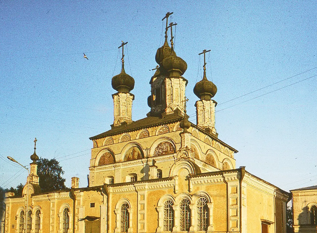 A yellow and white stone church with five dark onion domes and tall arched windows under a clear blue sky