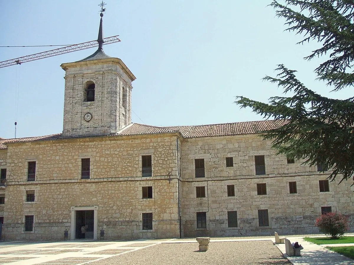 A long three-story stone monastery building with a square bell tower and a cedar tree, under a clear blue sky