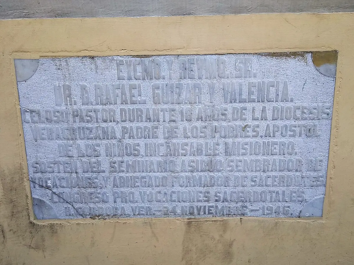 Weathered stone plaque on a yellow wall with carved Spanish capital letters praising Bishop Rafael Guízar y Valencia