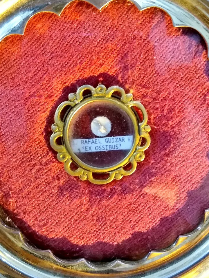 Small round gold reliquary on red velvet holding a tiny fragment, with a label reading Rafael Guízar y and ex ossibus