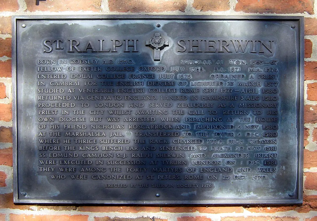 Dark metal plaque on a brick wall headed "St. Ralph Sherwin" with a cross and several lines of raised text about his life