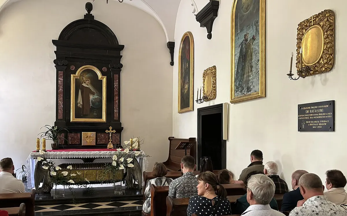 A white chapel with a black marble altar holding a painting of the saint, a reliquary below it, framed paintings on the walls and people seated in prayer