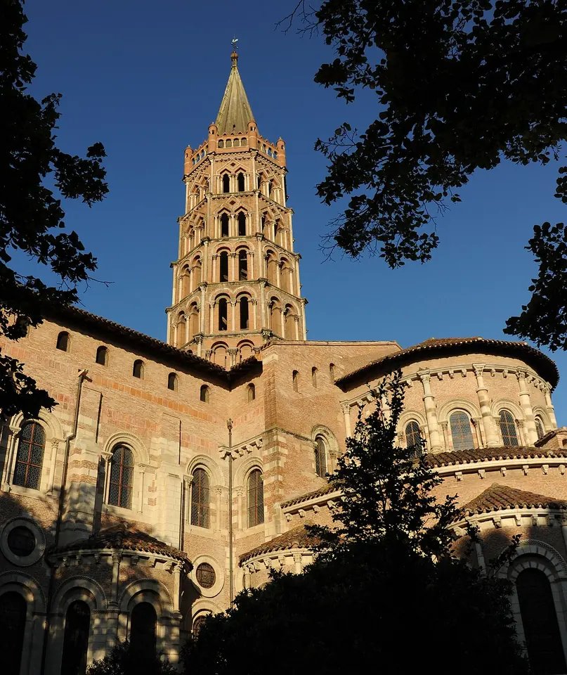 The rounded brick and stone chapels of a great Romanesque church rising in tiers to an octagonal bell tower with a spire, framed by dark tree branches against a blue sky