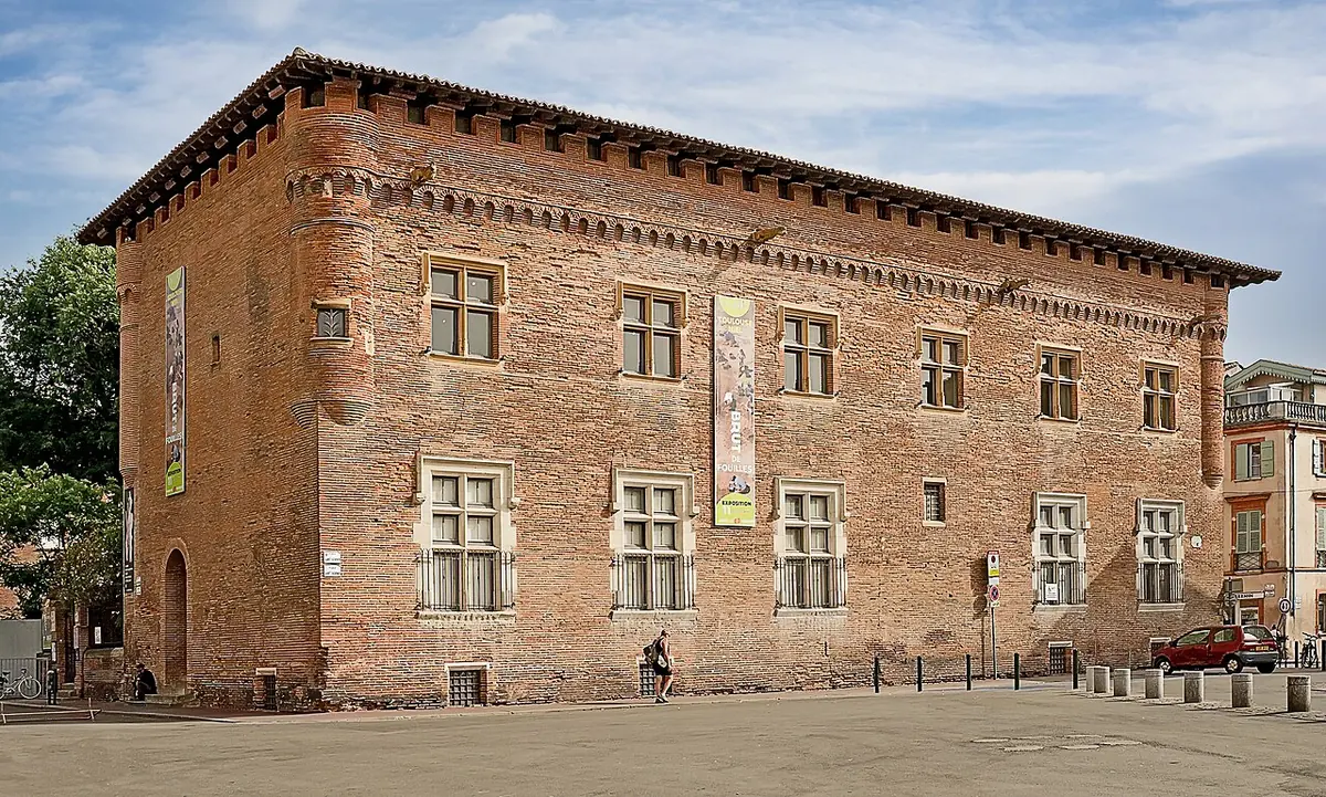 A long red-brick building of the sixteenth century with stone-framed windows and a battlemented roofline on a sunny square