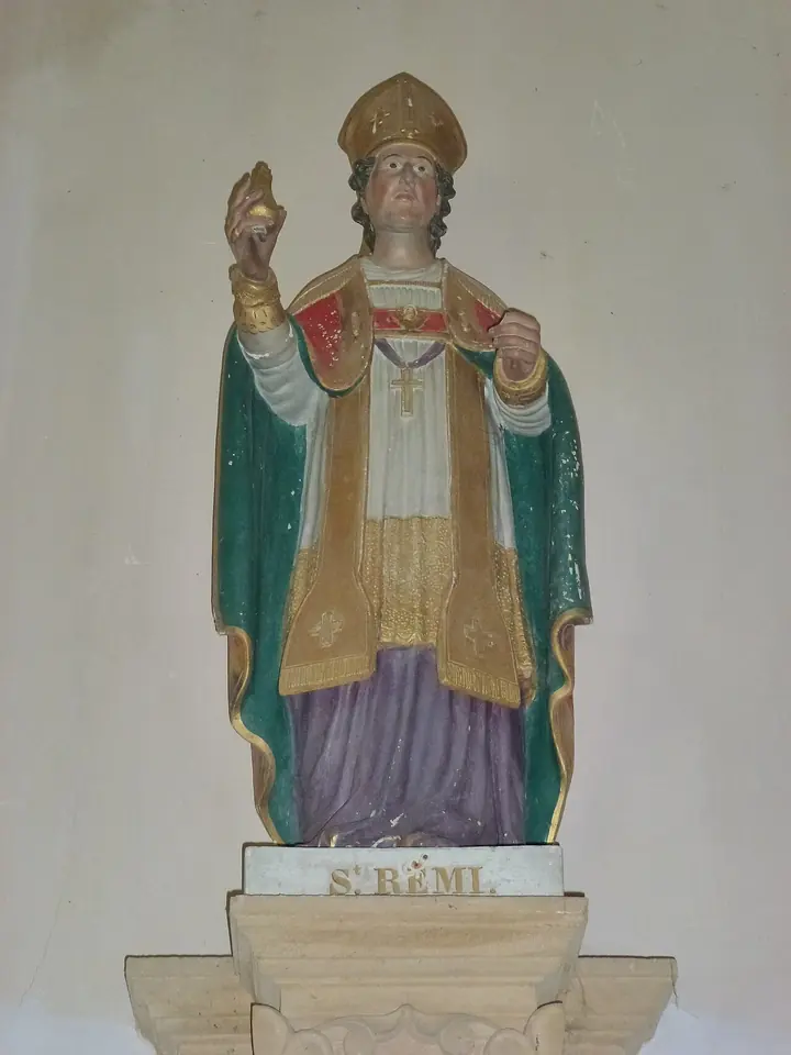 Painted statue of a bishop in a gold miter and green cope over red and white vestments, raising a small golden flask in his right hand, on a pedestal labeled St. Remi