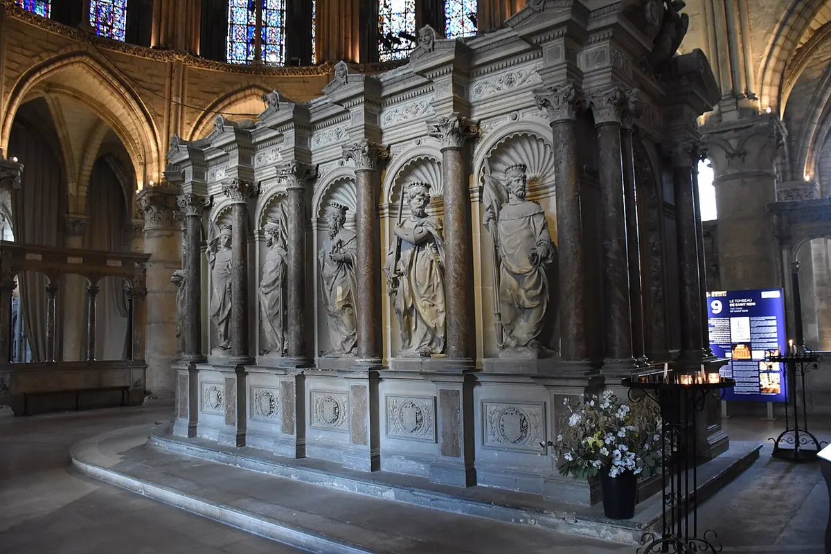 A large carved stone tomb monument with statues of bishops and nobles in arched niches, standing in the choir of a Gothic church with stained-glass windows above