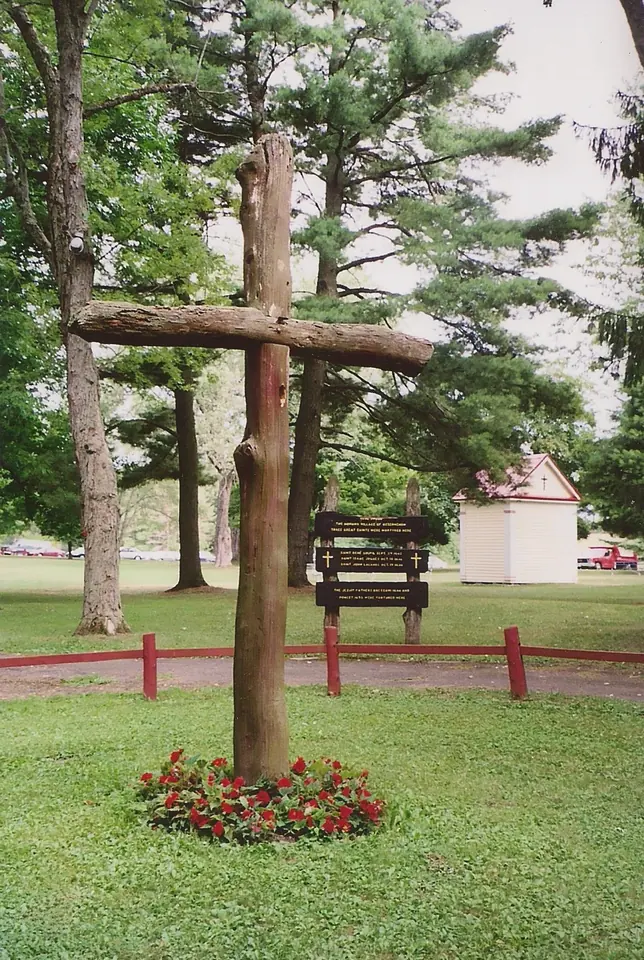 A tall rough-hewn wooden cross rising from a bed of red flowers among pine trees on the shrine grounds at Auriesville