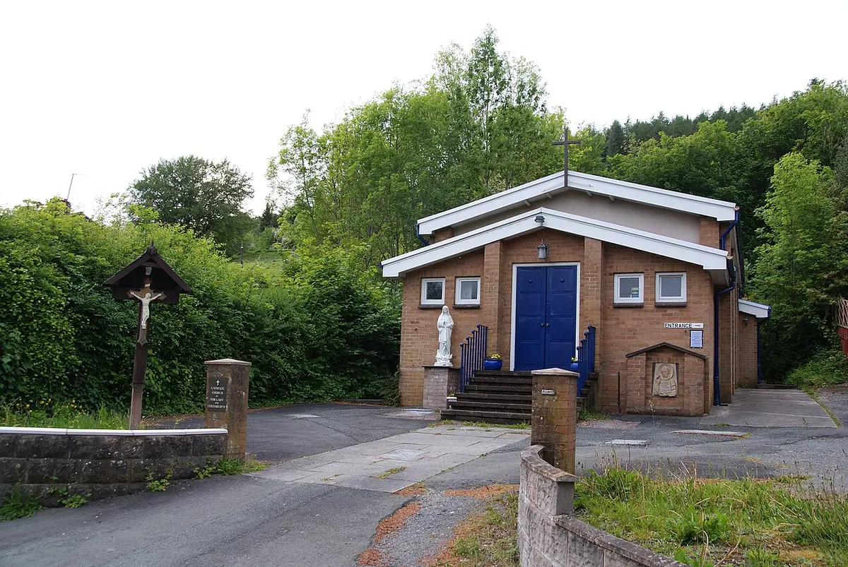 A small modern brick Catholic church with a blue door and a wayside crucifix, set among green trees