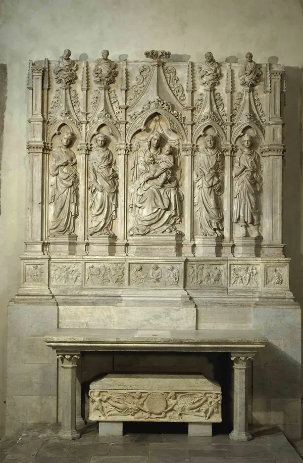 Carved marble altarpiece with the Virgin and Child among standing saints in Gothic niches, above an altar with a stone sarcophagus beneath it