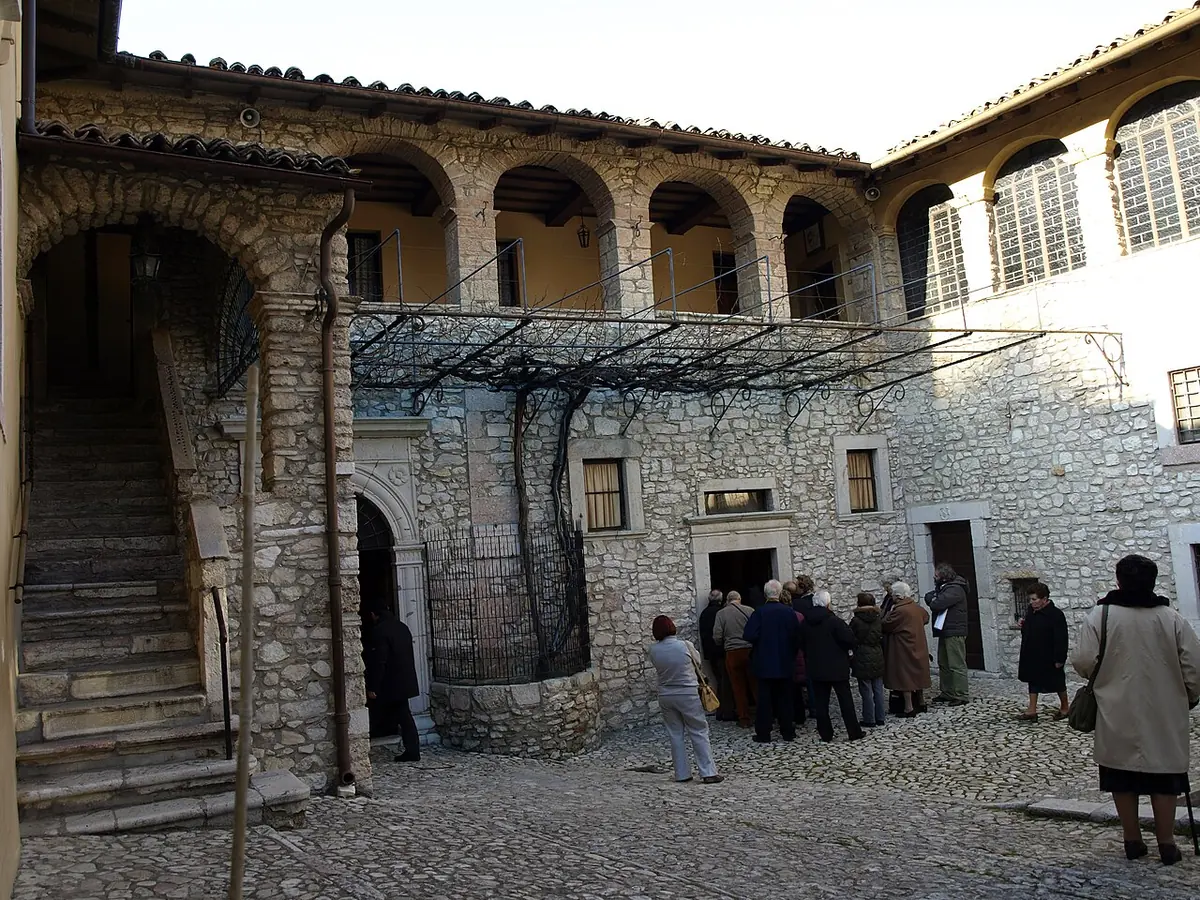 Pilgrims in a stone courtyard of the monastery of St. Rita in Cascia, with an arcaded upper gallery and a vine trellis