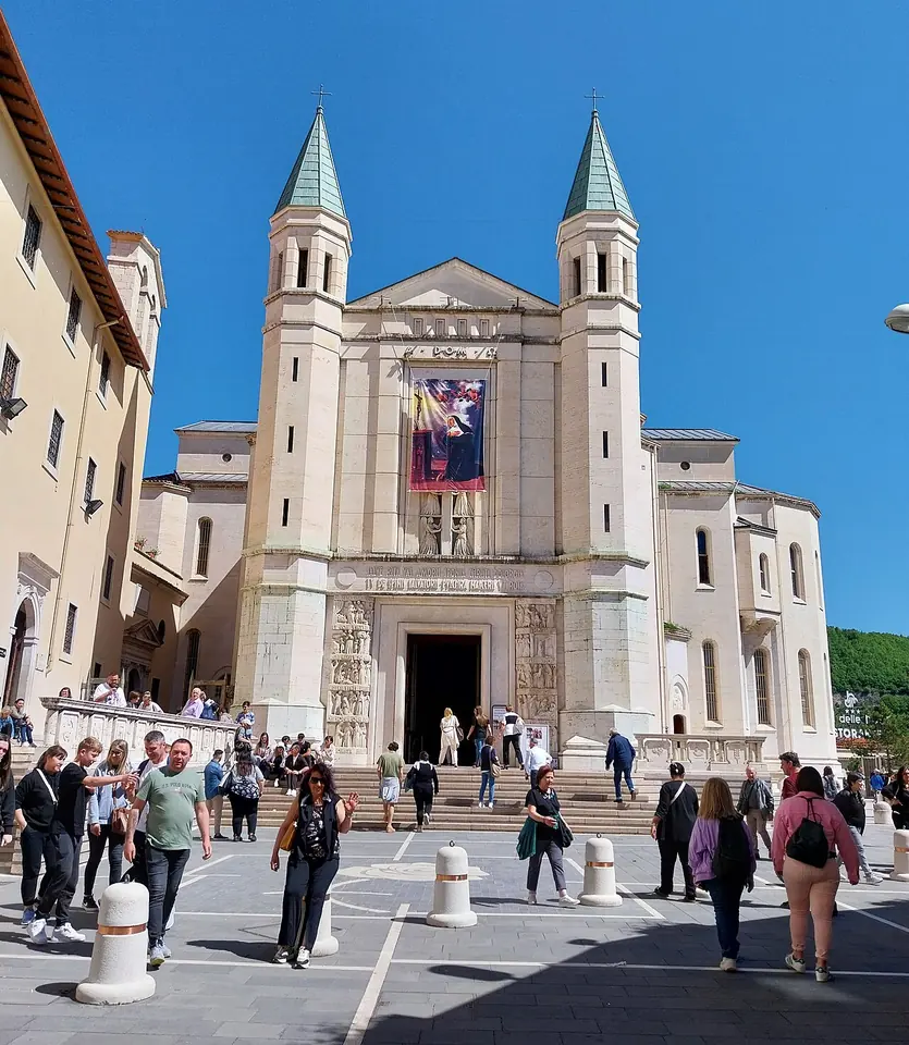 White travertine facade of the Basilica of Santa Rita with two green-capped bell towers, a banner of the saint and pilgrims on the steps
