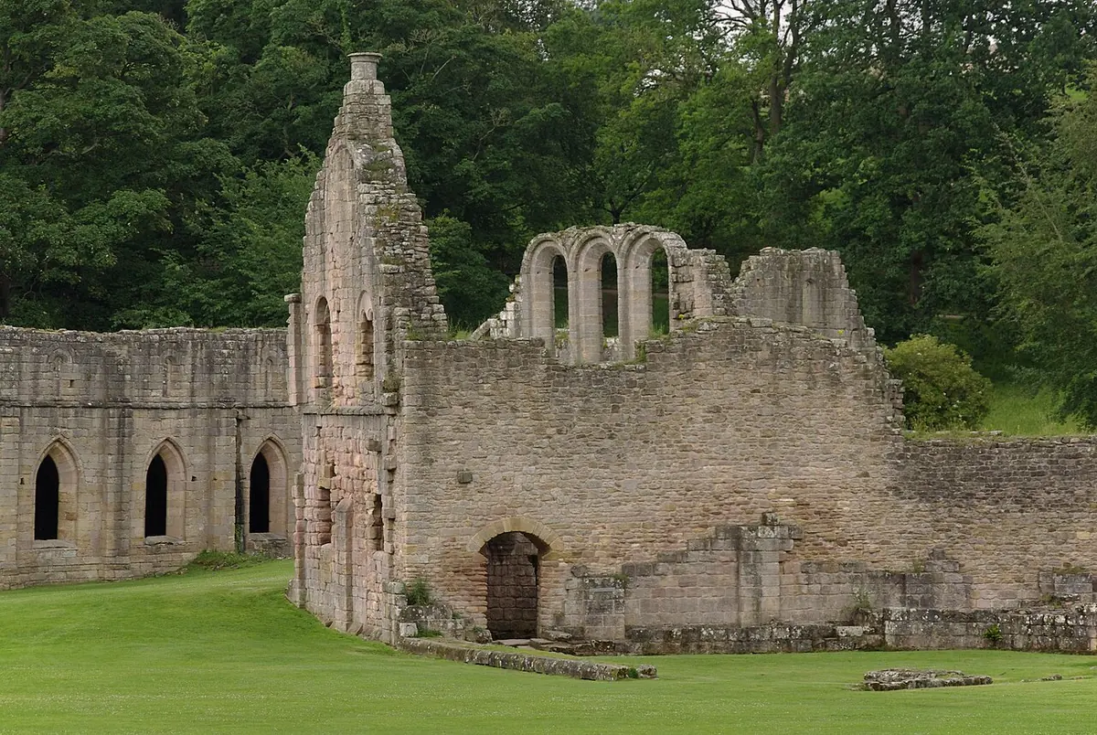 The roofless stone ruins of a great Cistercian abbey on a green lawn, with tall lancet windows and arcades against a wooded hillside