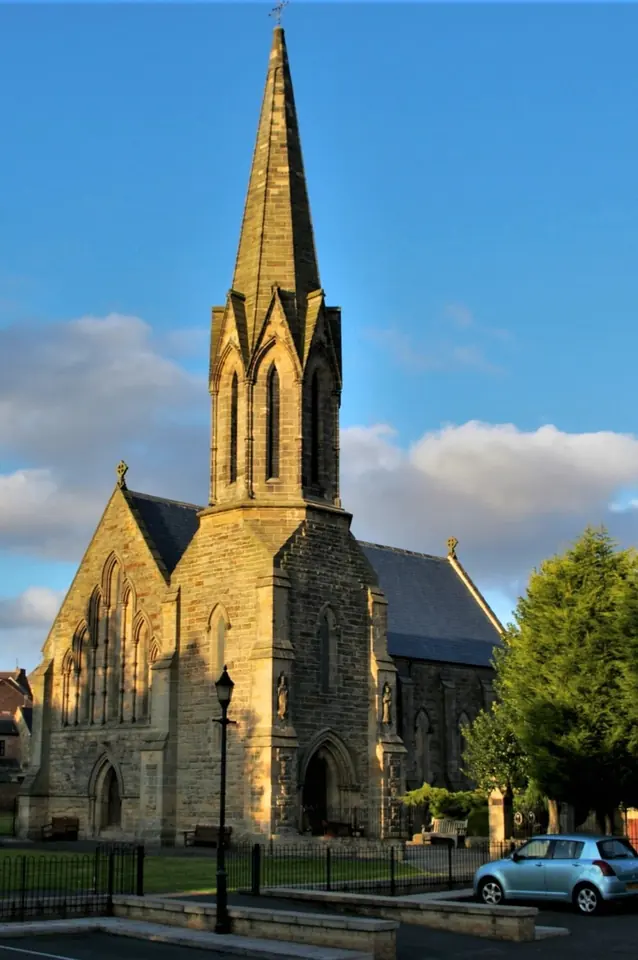 A sandstone Gothic Revival church with a tall spire, lit by low evening sun under a blue sky with a few clouds