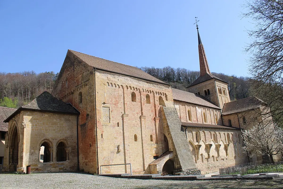 Romanesque stone abbey church with a tall pointed bell tower and an arcaded porch, set against wooded hills under a blue sky