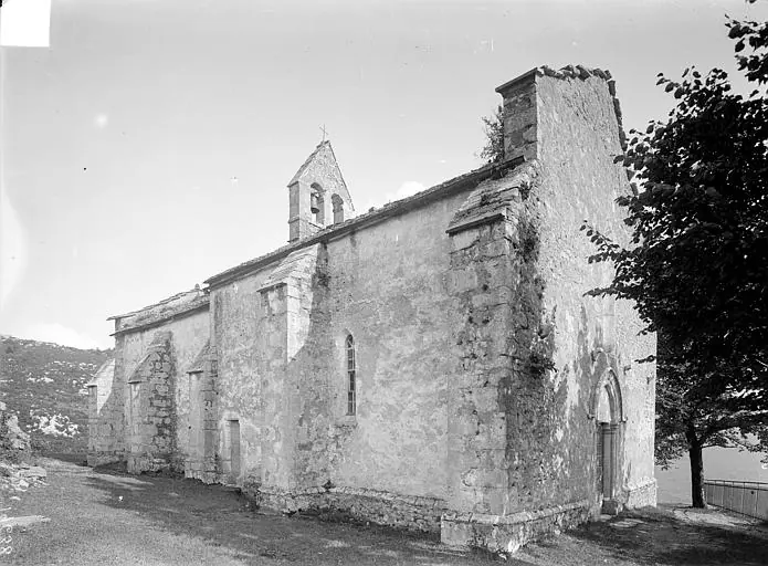 Old black-and-white photograph of a small stone country chapel with a low bell gable, standing on a rocky slope beside a tree