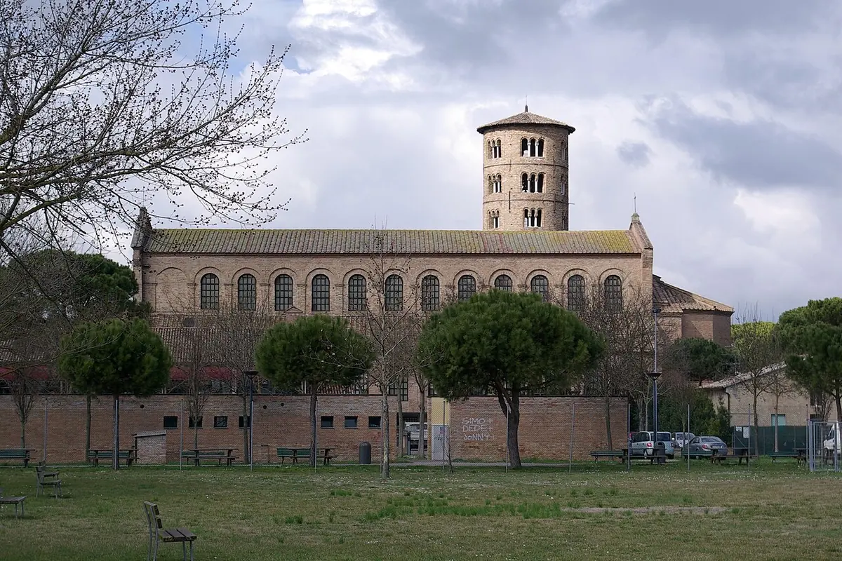 Long brick basilica with a tall round bell tower, seen across a lawn with pine trees near Ravenna
