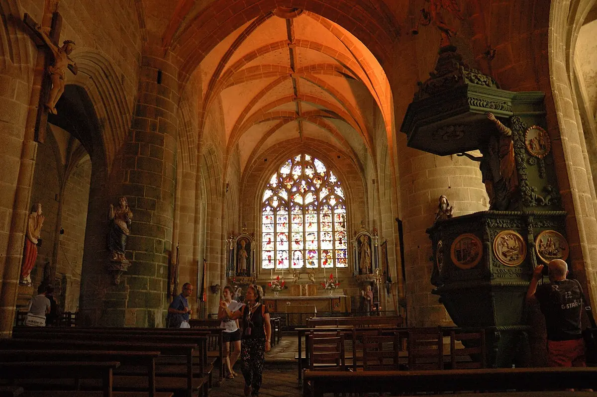 Interior of a granite Gothic church with a tall stained-glass east window, wooden pews and a green carved pulpit with painted medallions on the right