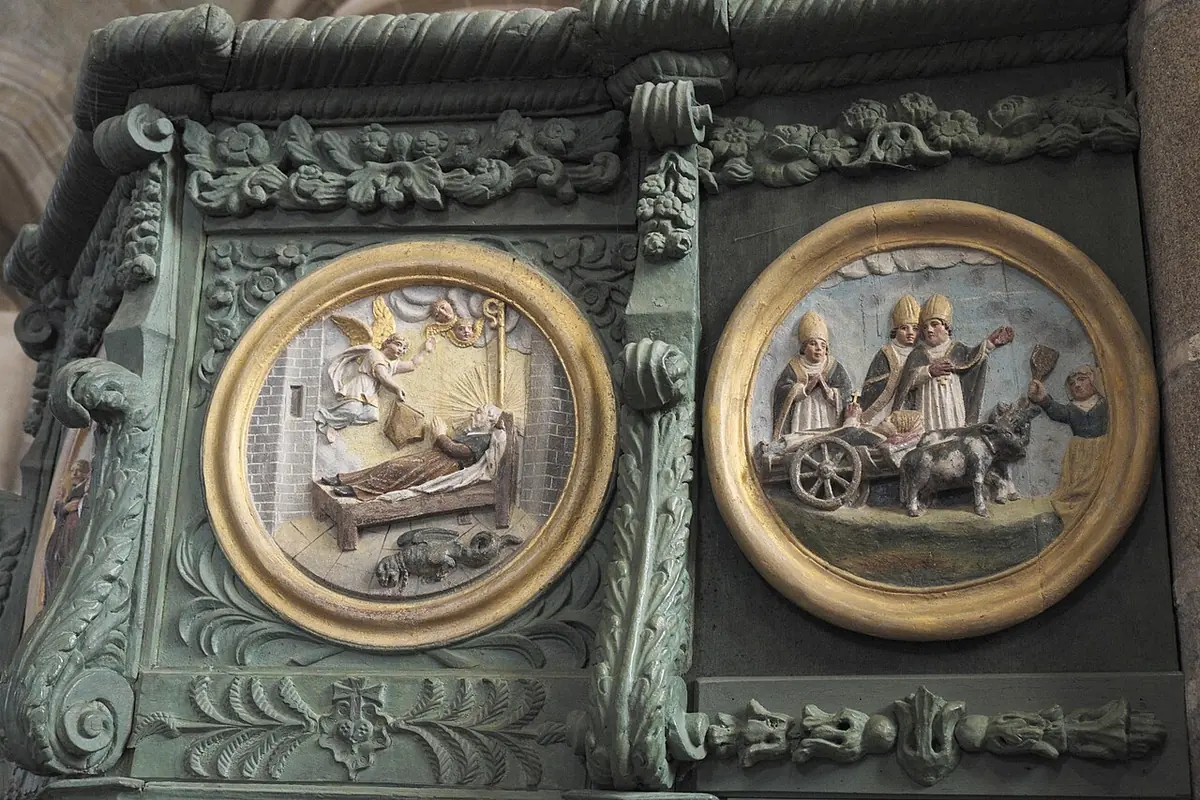 Two painted round medallions on a green carved wooden pulpit, one showing a monk on his deathbed and the other a cart drawn by oxen carrying his body with figures beside it