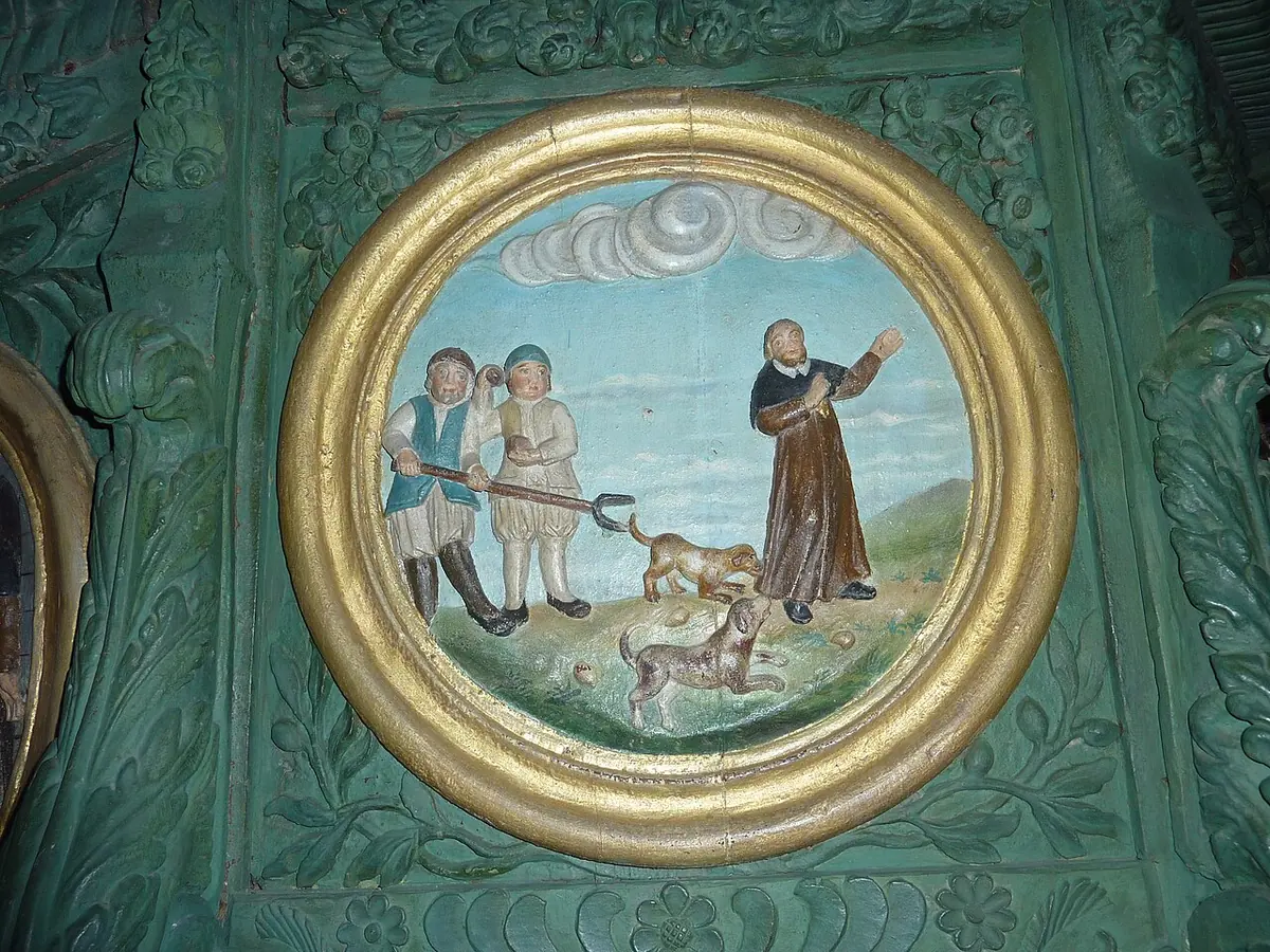 Painted round medallion on a green carved wooden pulpit showing a monk in a brown habit blessing two hounds while two men watch