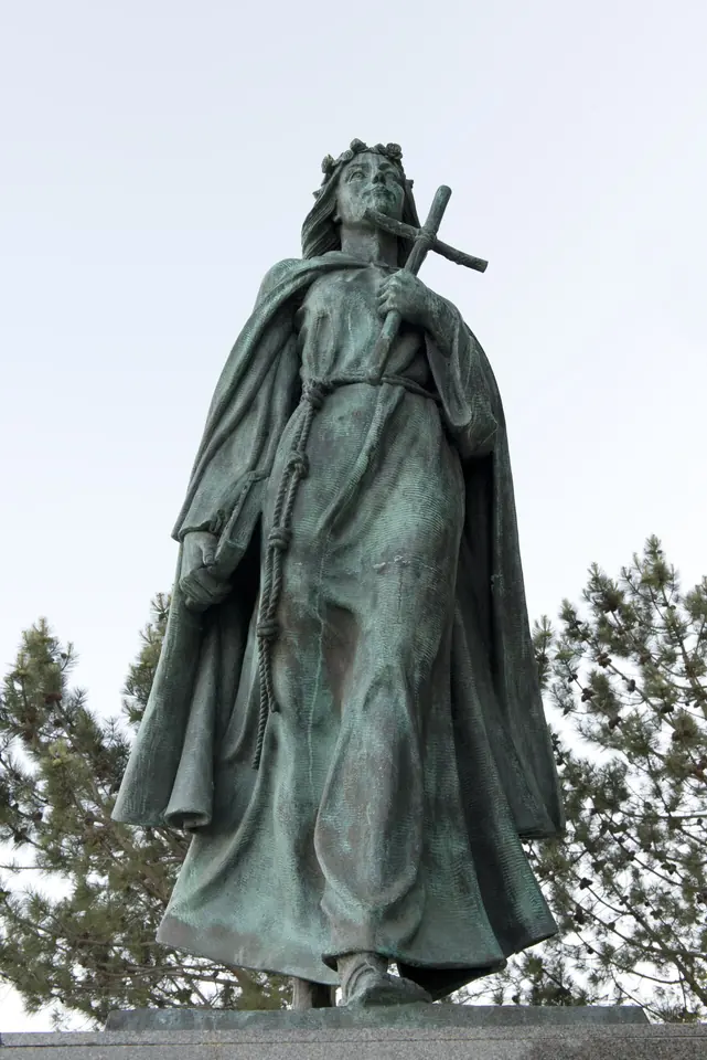A weathered green bronze statue of St. Rosalia in a hooded cloak holding a cross, standing against a pale sky in Monterey, California
