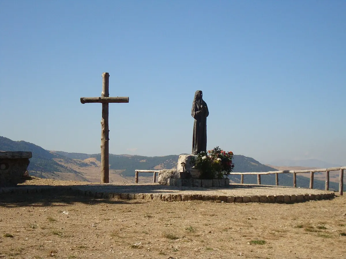 A dark statue of St. Rosalia beside a tall wooden cross on a rocky hilltop, with rolling Sicilian hills behind