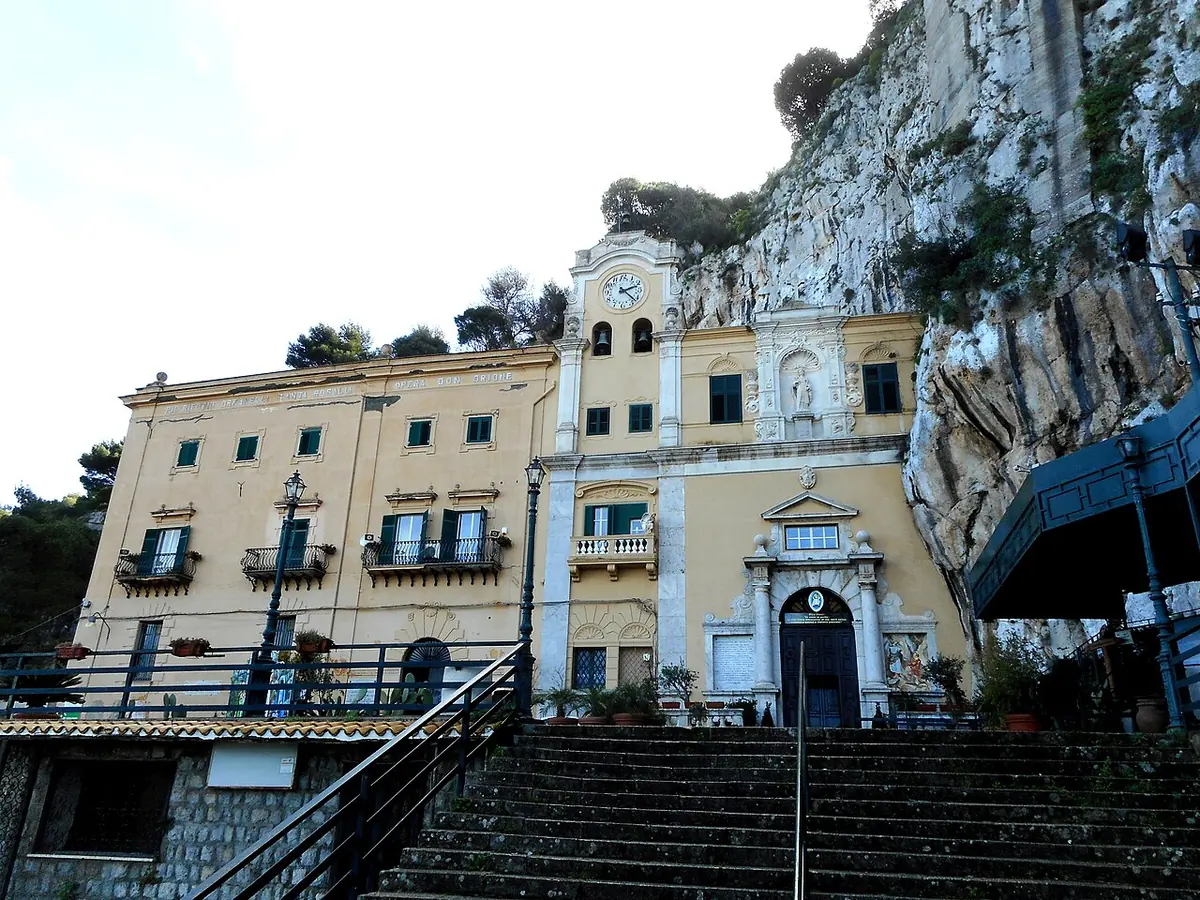 The yellow 17th-century facade of the Sanctuary of St. Rosalia, with a clock tower, built against a sheer limestone cliff and reached by a flight of steps