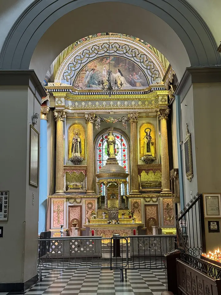 Gilded altar in a side aisle of a church, with statues of saints in niches and a glass reliquary at its center, behind a low railing