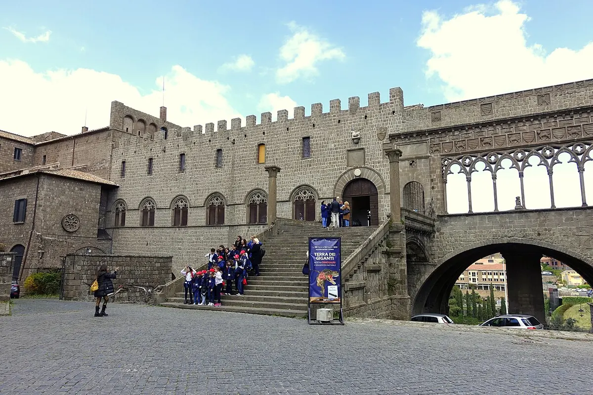 Medieval stone palace with battlements, a broad outdoor staircase and an open Gothic loggia on arches, with schoolchildren gathered on the steps