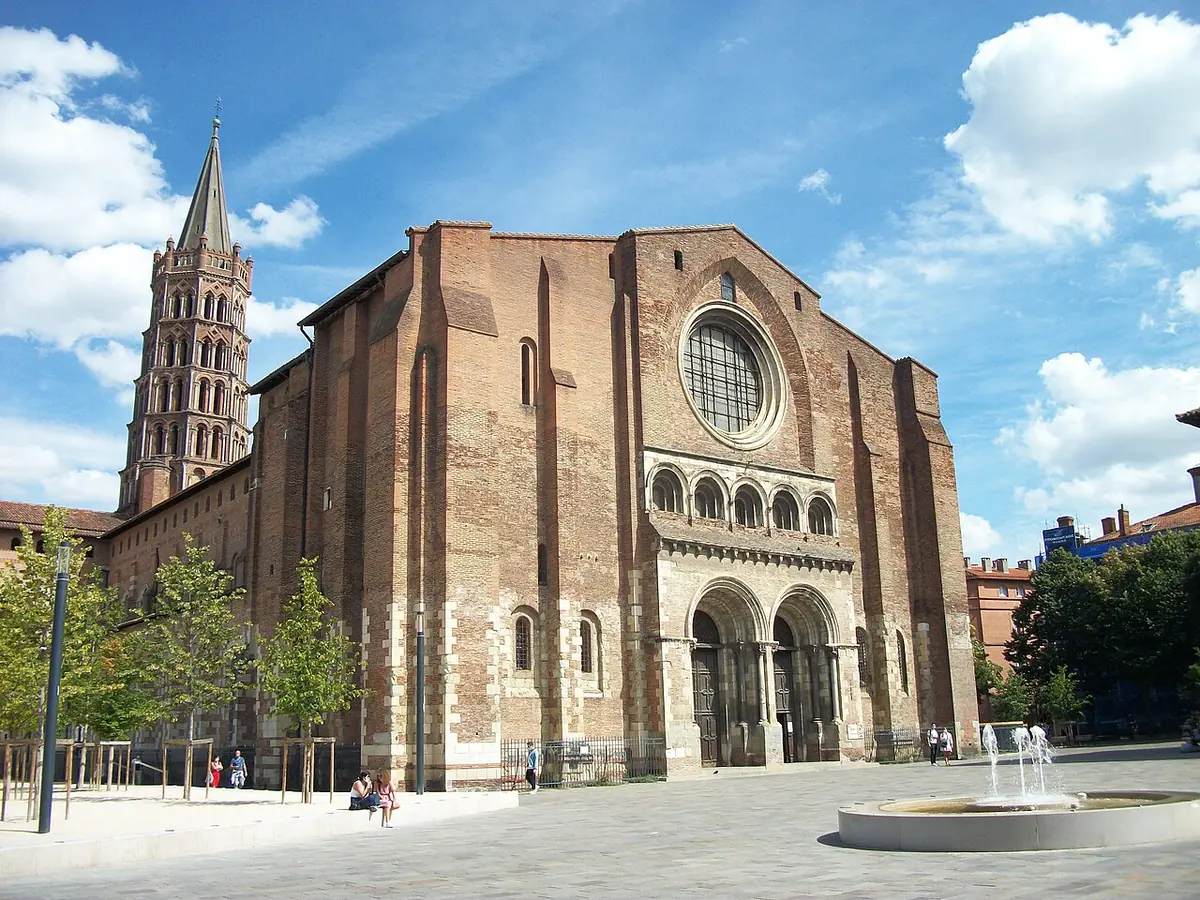 The long red-brick west front of a Romanesque basilica with a tall octagonal bell tower rising behind, seen across a sunny square