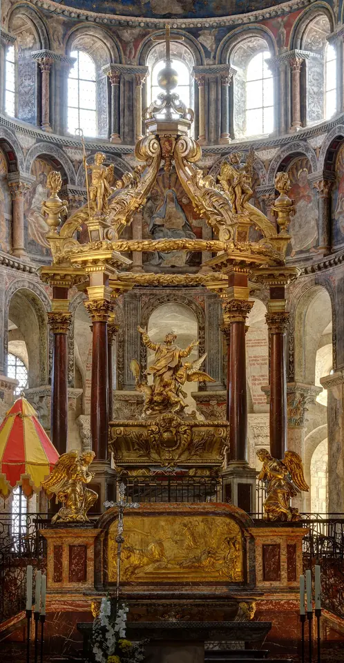 A tall gilded baroque canopy on red marble columns rising over a tomb in the apse of a Romanesque church, with a golden relief of a bull below