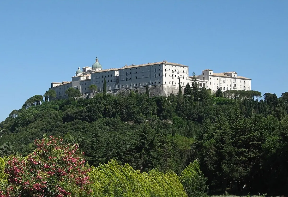 The long white buildings of Monte Cassino Abbey on top of a wooded mountain under a blue sky