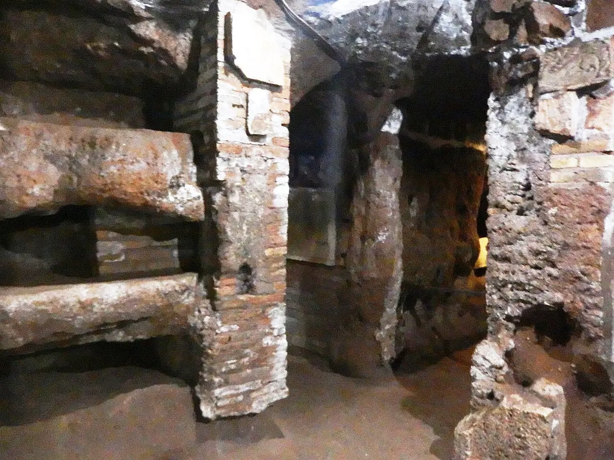 Narrow underground gallery cut into brown tufa rock, with rows of empty rectangular burial niches in the walls