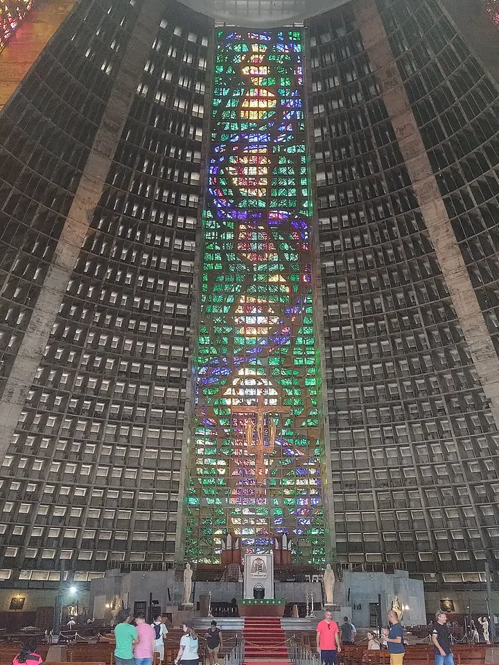 Interior of a vast cone-shaped concrete cathedral with a tall stained-glass window rising to the roof above the altar and rows of pews