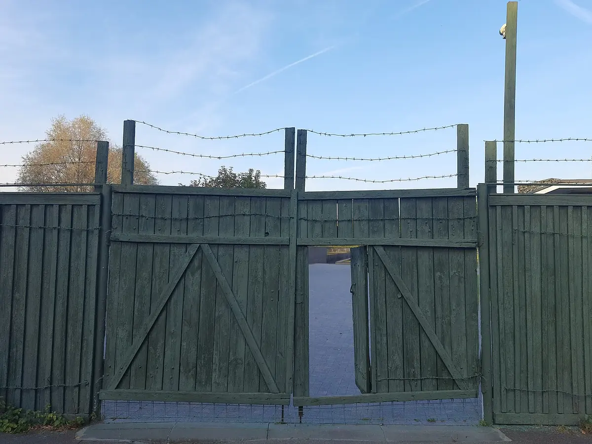 High green wooden fence and gate topped with barbed wire under an autumn sky, with bare trees beyond