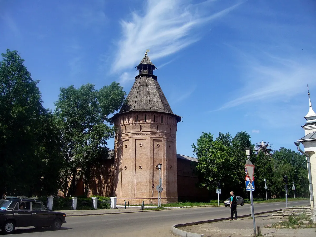 Massive red-brick monastery wall with a tall tower under a tent roof, trees and a quiet road in front