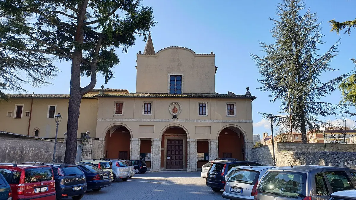 Plain cream-colored friary church with a small bell tower above an arched portico, with tall cedars and parked cars in front