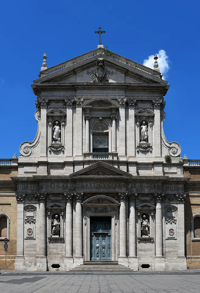 Two-story travertine church facade with columns, statues in niches and a triangular pediment topped by a cross, under a blue sky