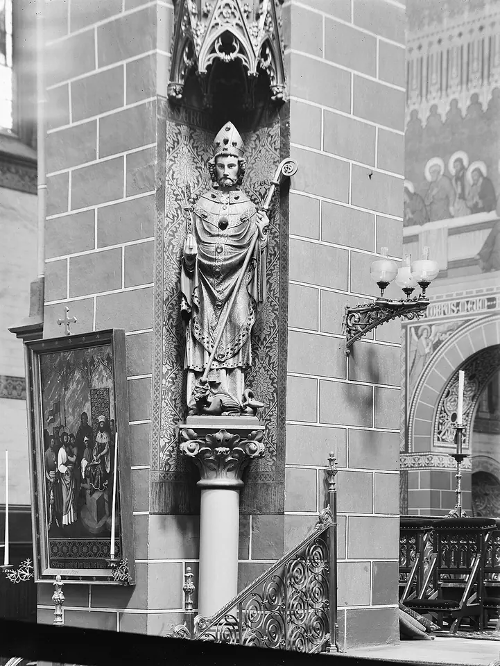 Old black-and-white photograph of a carved statue of a mitered bishop with a crozier on a bracket against a church pillar