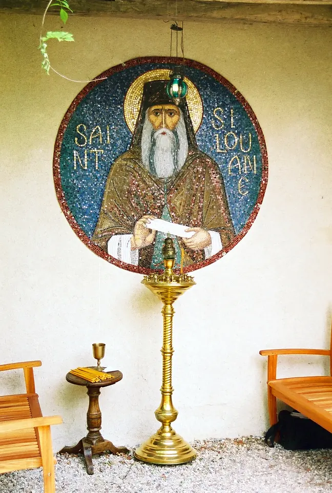 Round gold mosaic of St. Silouan with a gray beard and monastic hood, holding an unrolled scroll, above a tall brass oil lamp stand on a whitewashed porch