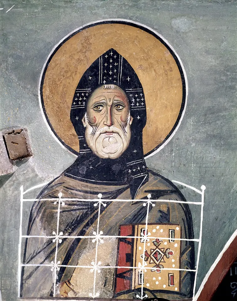 Byzantine fresco of an old monk with a long white beard and a dark cowl marked with white crosses, standing above a column with a railed top and holding a book