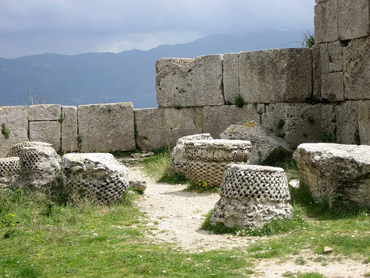 Ruined limestone walls and fallen carved stone blocks in grass on a hilltop, with mountains under clouds in the distance