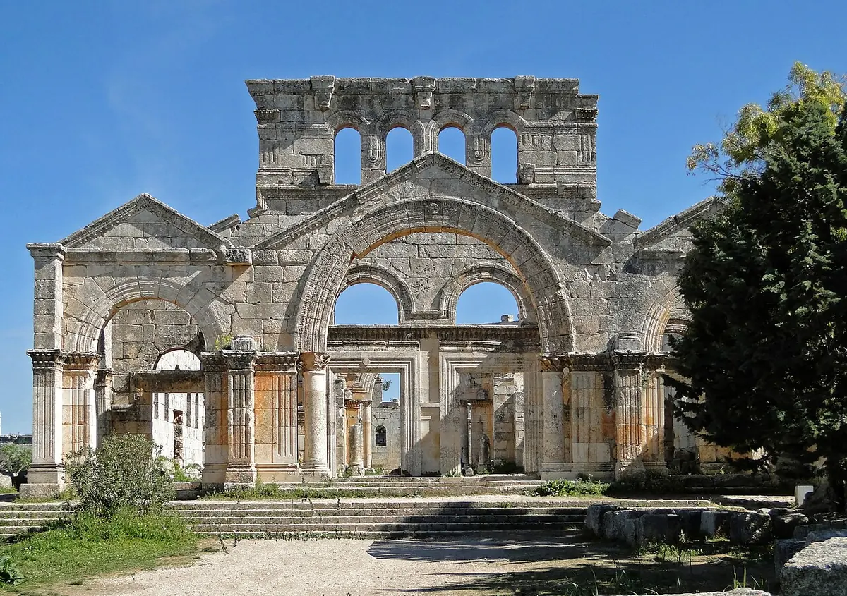 Roofless 5th-century stone church facade with a great central arch, smaller arches and tall arched windows under a blue sky, with grass and a tree in front