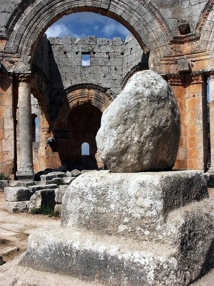 A rounded, worn stone block sitting on a square stone base in the middle of ruined arches, all that was left of Simeon's pillar in 2005