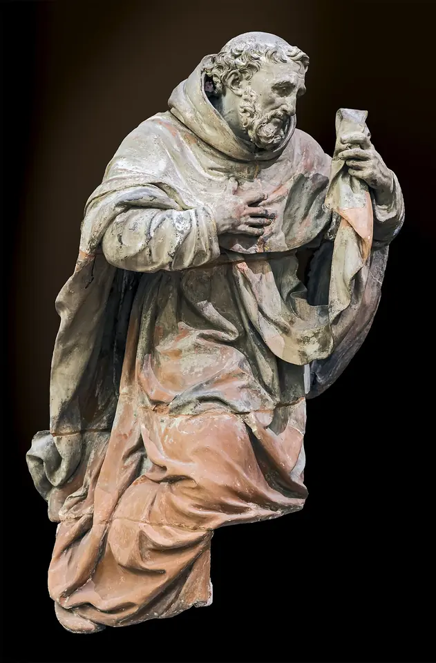 Terracotta statue of an old bearded friar in a heavy cloak, one hand pressed to his chest and the other holding up a small scapular, his face lifted toward heaven