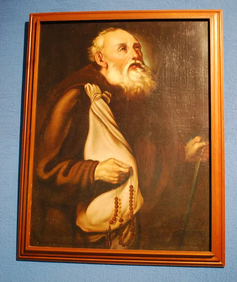 Painting of an old bald friar with a long white beard in a brown habit, looking upward and holding a large white scapular and a rosary, with a walking staff